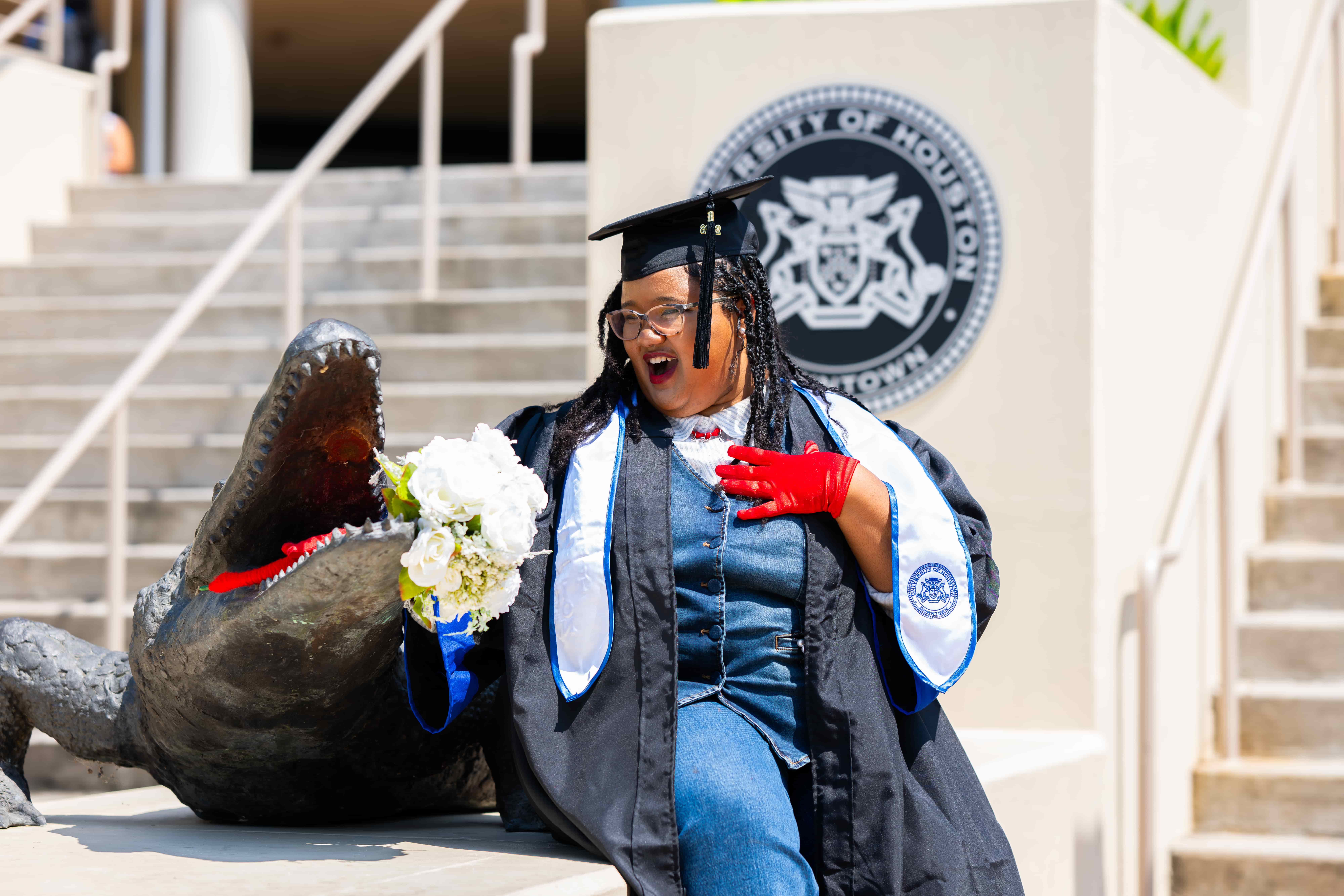 female graduate with red gloves in her cap and gown next to the gator statue at mdcob