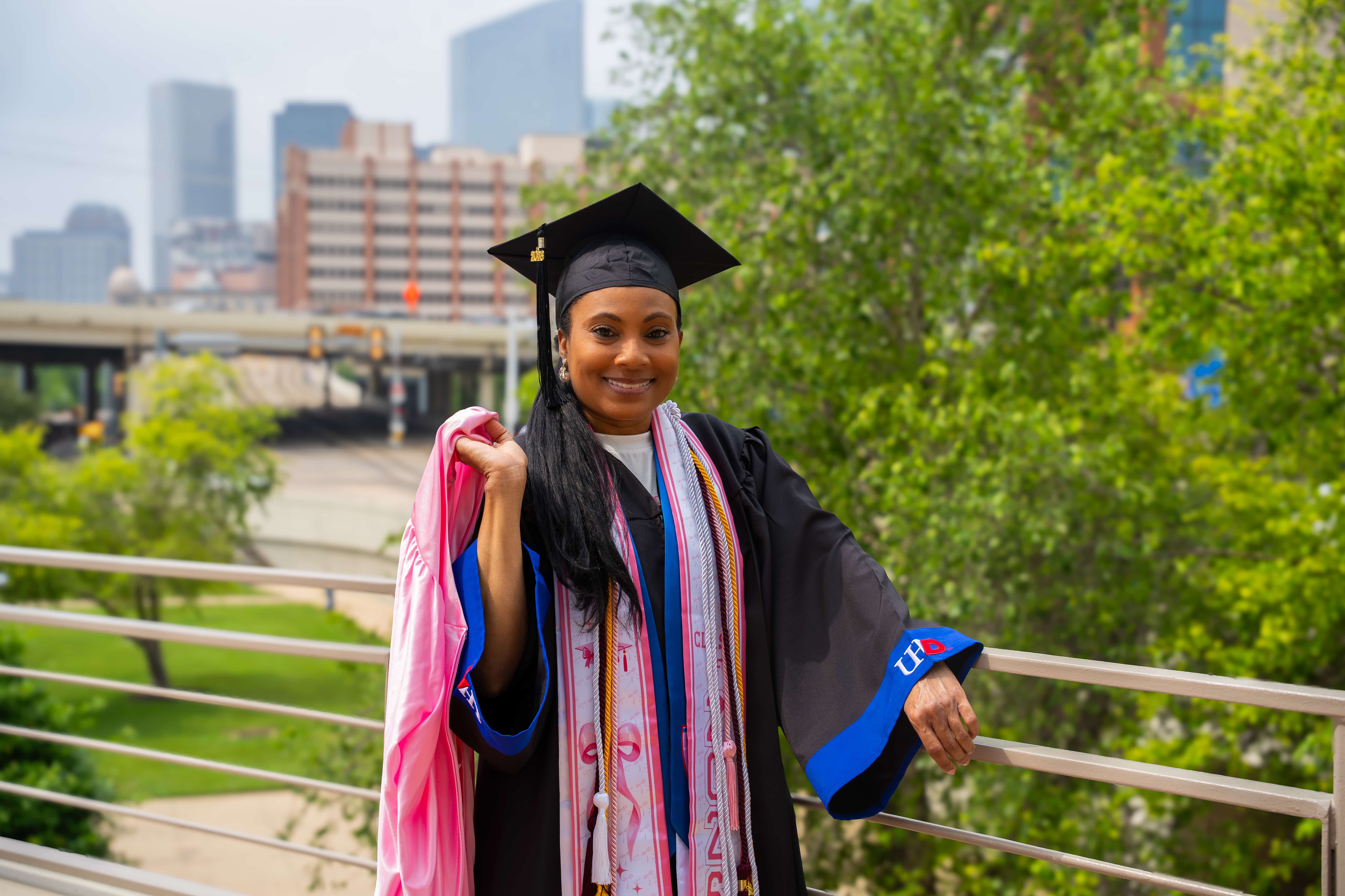 student in her UHD grad cap and gown with a stole covered in pink bows holding a pink grad gown over her shoulder in the background, the city scape and a large tree