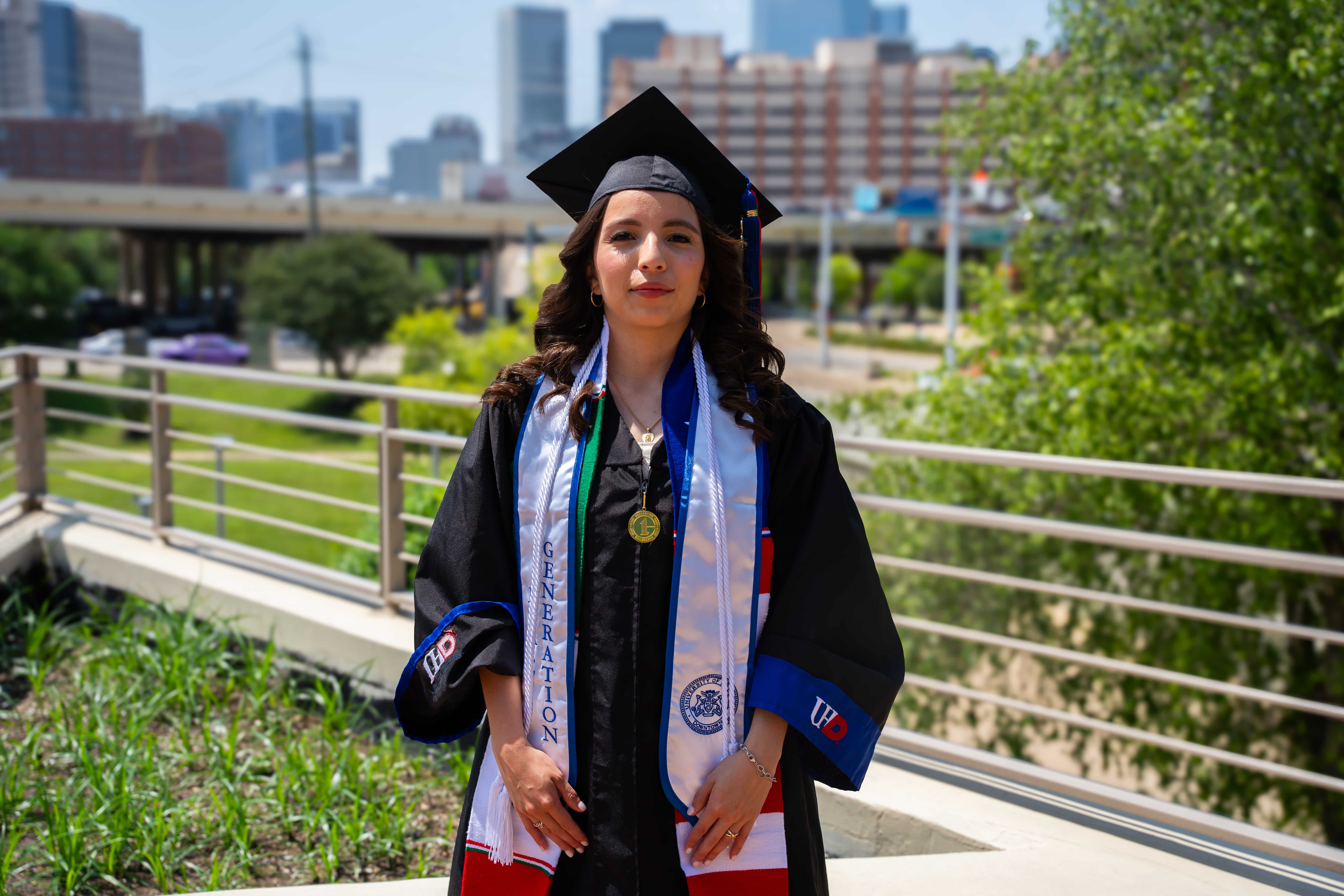 female uhd graduate in cap and gown with a cityscape background