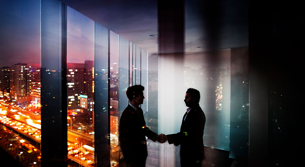 Two men shaking hands near a window at night, with a skyline in the background.