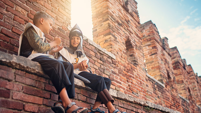 Two kids wearing armor and sitting on the wall of a castle
