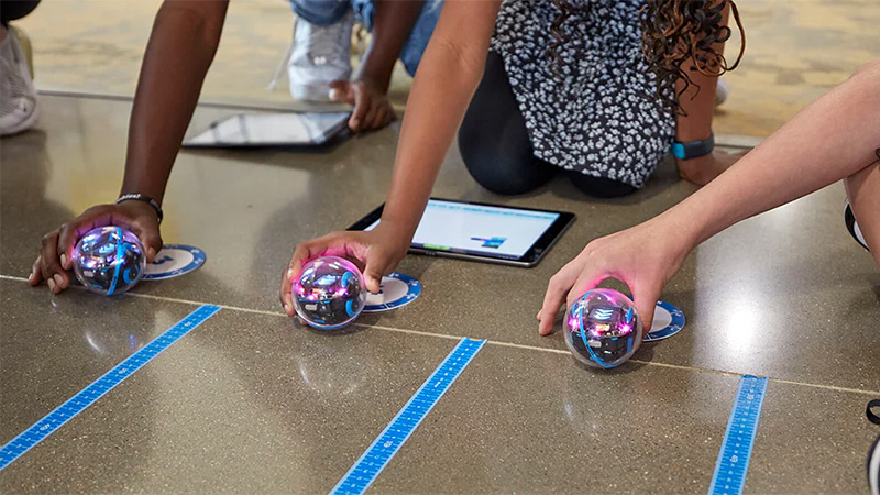 Hands of three teens holding Sphero robots on the floor