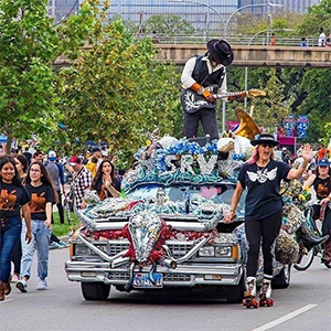 Art car and parade with made playing a guitar and woman roller skating beside the car