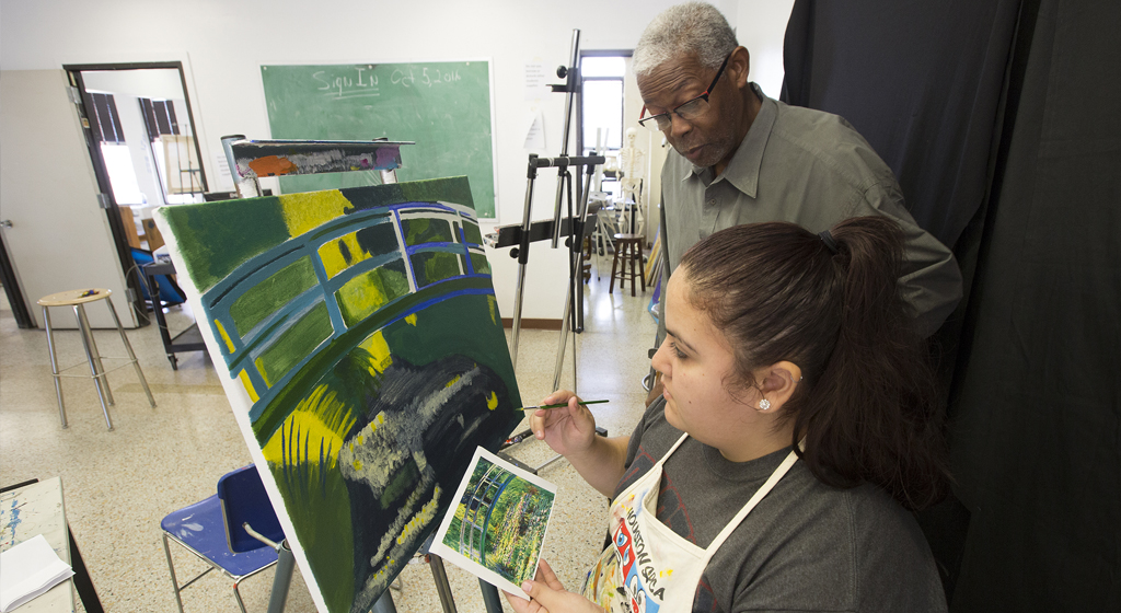 UHD Art professor Floyd Newsum instructs a student on painting at an easel.