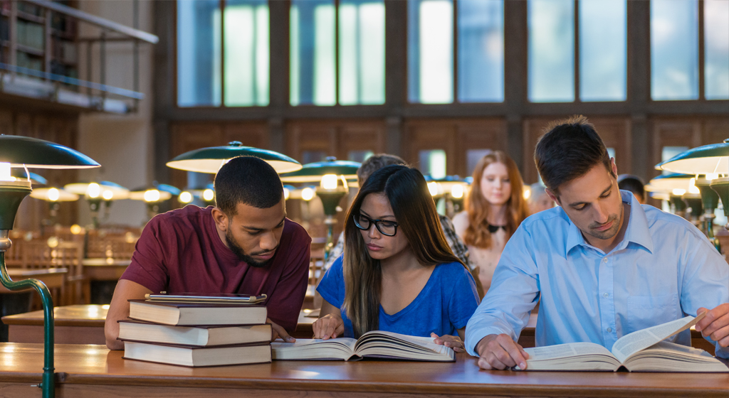 three students at a table in a library reading the same book doing research.