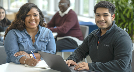 female student writing in a notebook at a table with a male student on a laptop in the UHD Welcome Center