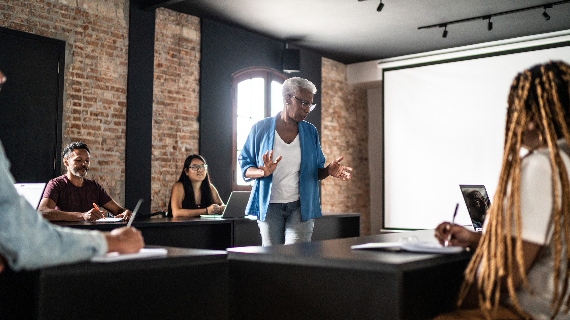 female professor stands between desks of students, giving a lecture