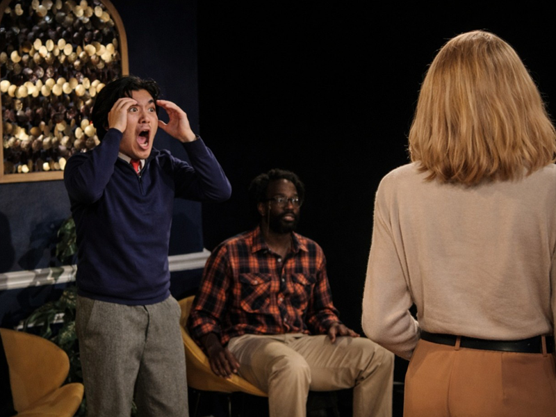 shocked male actor with hands on head looks at female actor while another male actor is seated on a couch