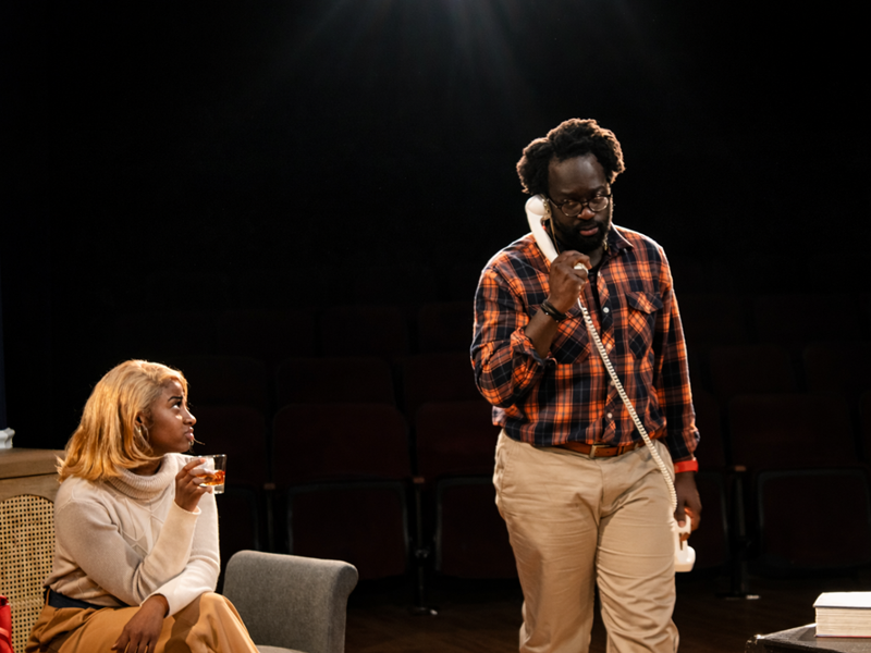 actress seated on sofa while male actor is on a land line phone