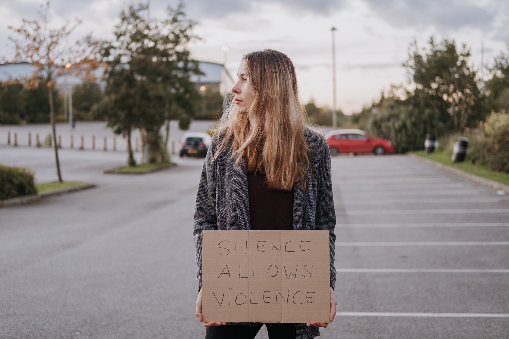 woman standing in a parking lot holding a cardboard sign that read 'silience is violence"