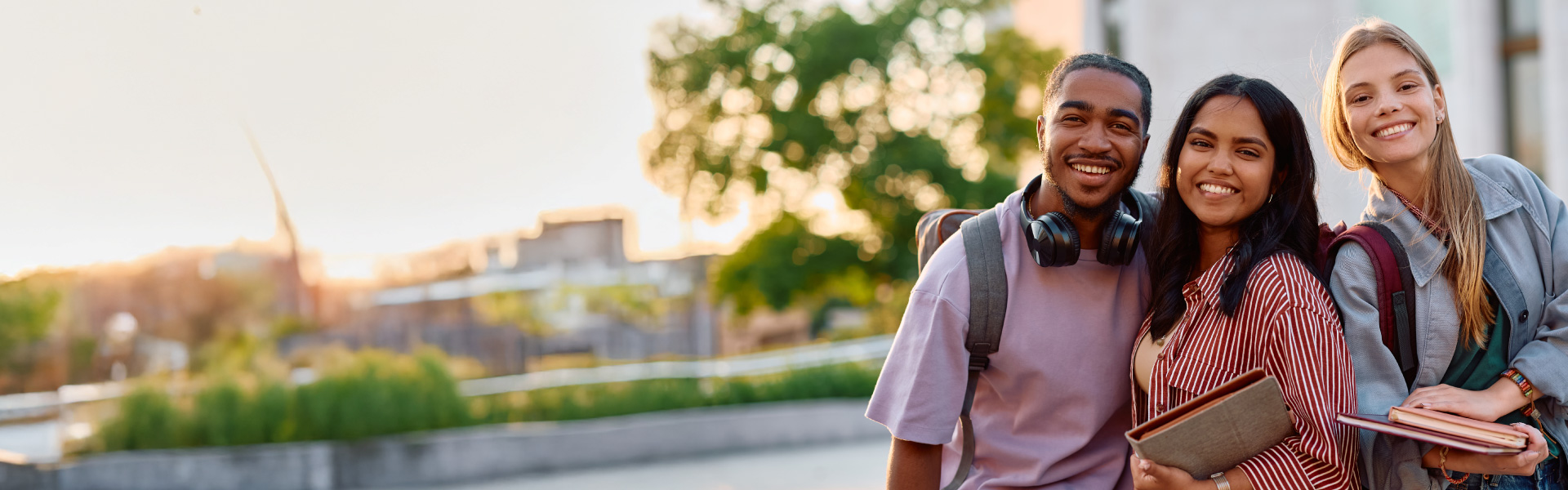Three happy students holding books and backpacks on a university campus, representing student life and course registration.