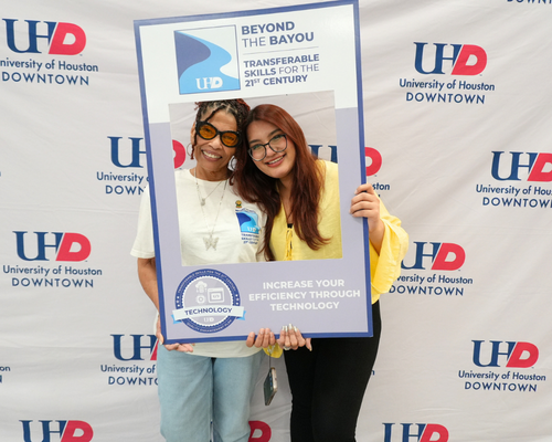Two students in University of Houston-Downtown's Bayou's Best smile and hold up a Quality Enhancement Plan frame in front of a step and repeat with the university's logo at the QEP Pep Rally.