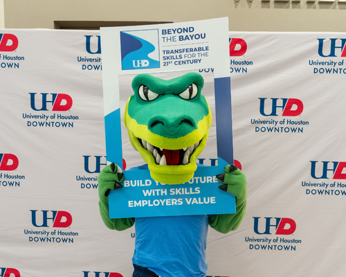 mascot for university of houston downtown edugator holds a frame in front a photo backdrop