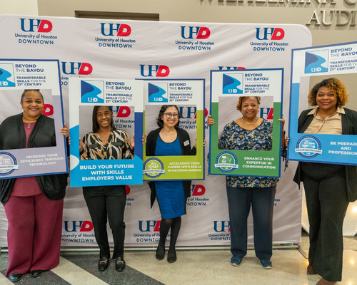 University of Houston-Downtown staff smile in front of a step and repeat while holding frames with the quality enhancement plan logo and text which reads beyond the bayou, transferable skills for the 21st century