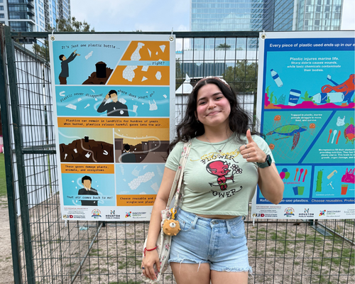 A University of Houston-Downtown student poses next to graphic design posters created in an Arts and Communication course led by Professor Poggio