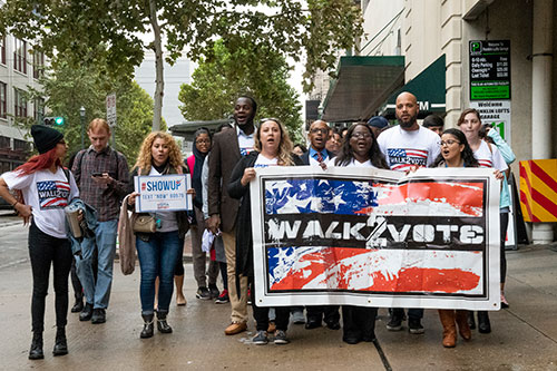 A group from the UHD community walking on the sidewalk with a flag that says walk to vote.