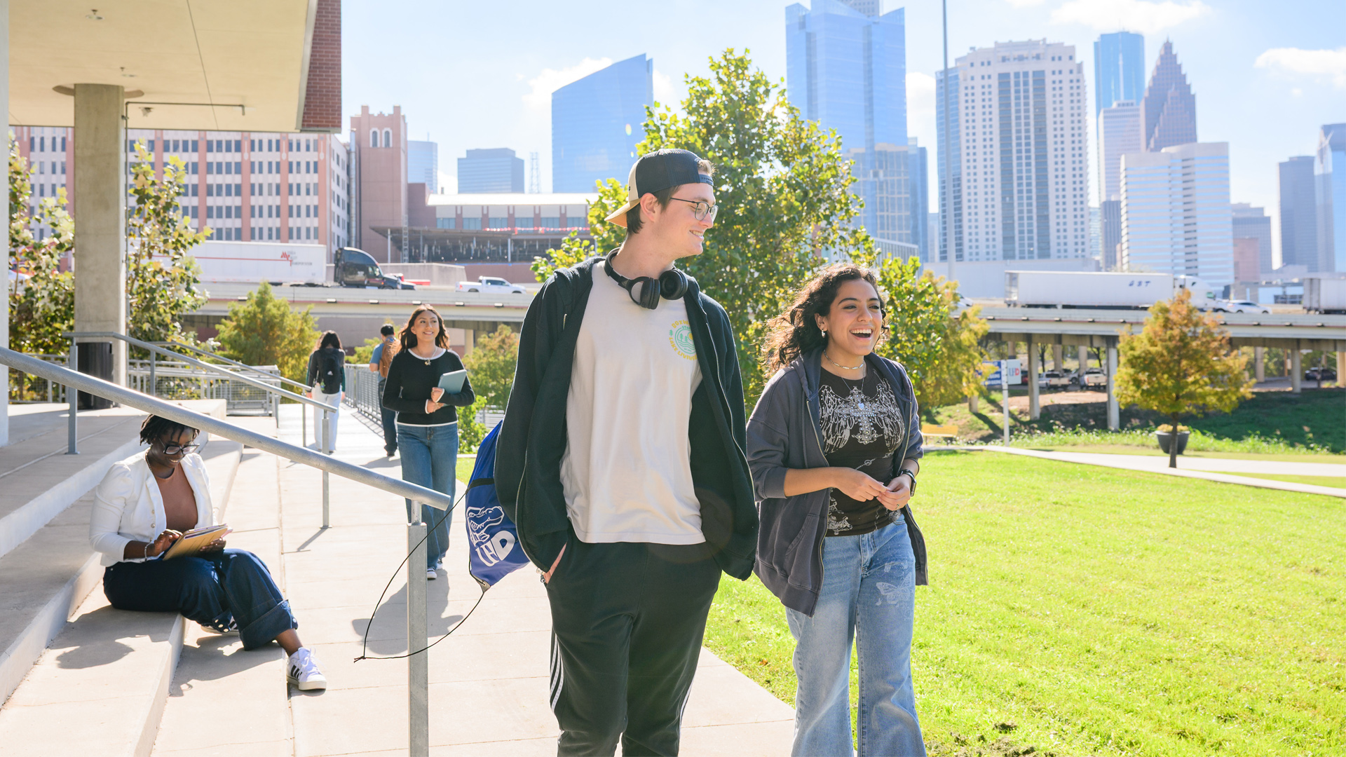 two UHD students walking outside in front of a campus building with Houston downtown buildings in the background