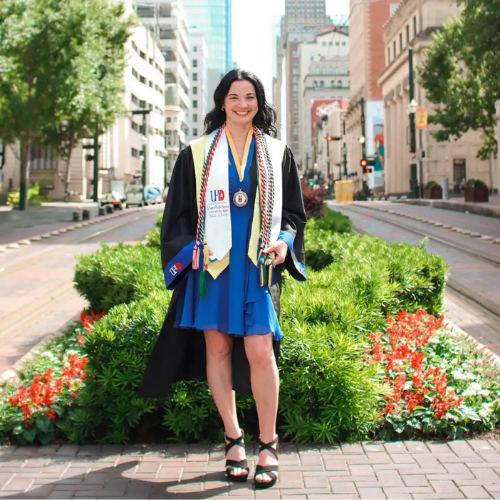 Social Work grad Amber Perez wears cords, stole, and graduation gown with University of Houston Downtown logo while smiling on Main Street in downtown Houston