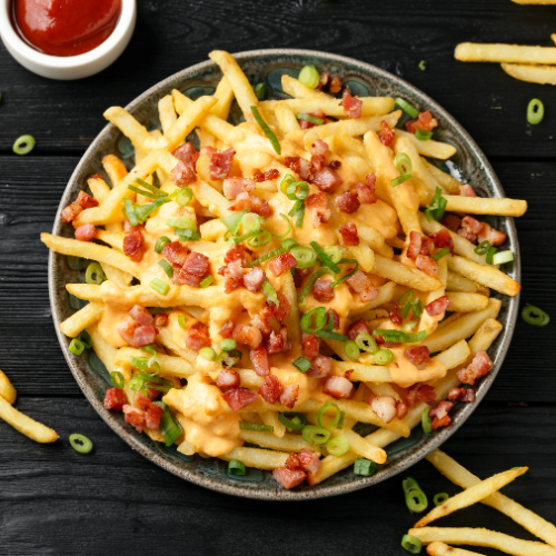 A bowl of loaded fries with bacon bits, chives, and cheese on a table with a small bowl of ketchup and stray french fries