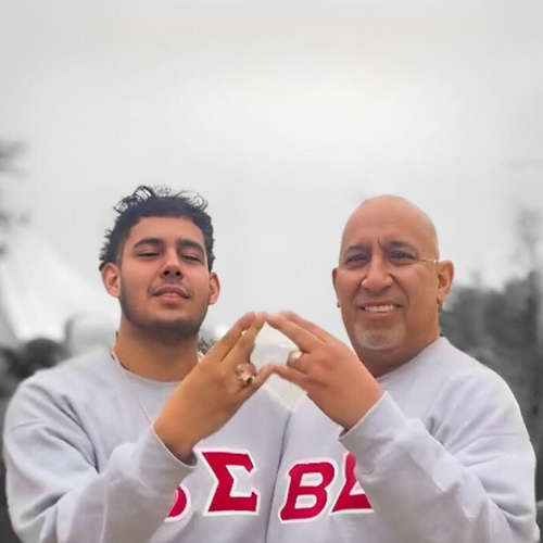 Armando and Sebastian Rodriguez hold up fraternity hand signs while wearing Omega Delta Phi Fraternity sweatshirts. The father and son are university of houston-downtown alums.
