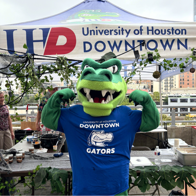university of houston-downtown mascot ed u gator holds their arms up in front of the university relations chili cook off tent at the university's annual chili cook off competition 