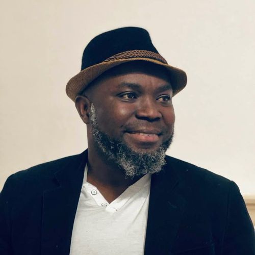 Photo of Godwin Agboka wearing a black hat, white shirt, and black sportcoat.