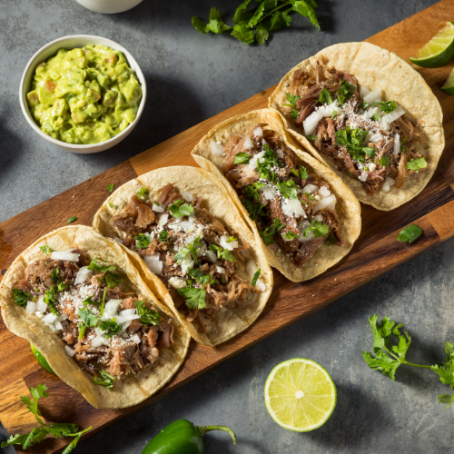 Four steak street tacos with cilantro and onion on a cutting board next to guacamole and a lime